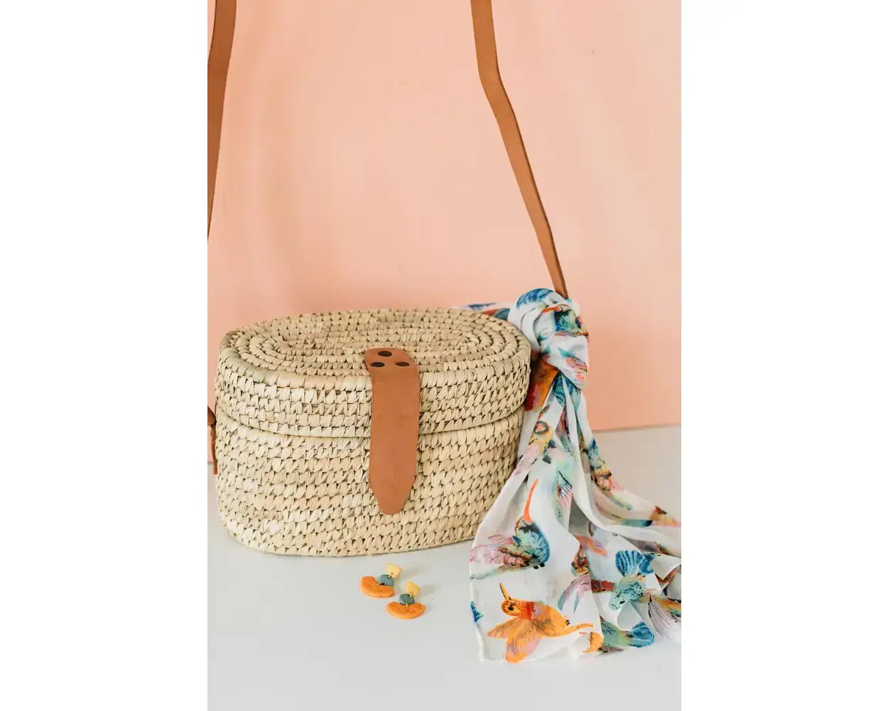 A delicate chiffon scarf with a floral and hummingbird design by Ten Thousand Villages, elegantly tied around a woven bag.