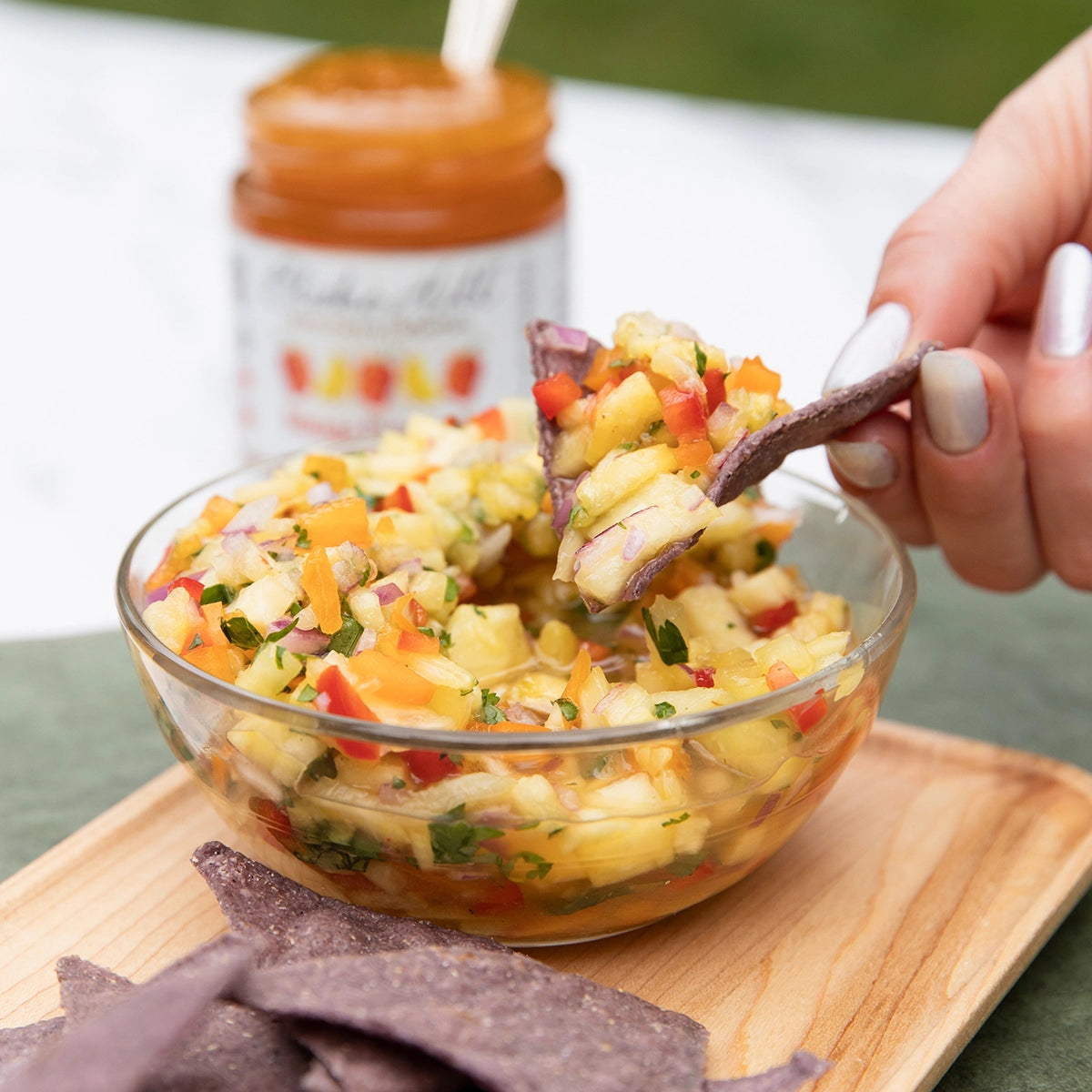 Glass bowl of colorful salsa made with Mango Habanero Jam by Blake Hill Preserves with a hand holding a tortilla chip, on a wooden board.
