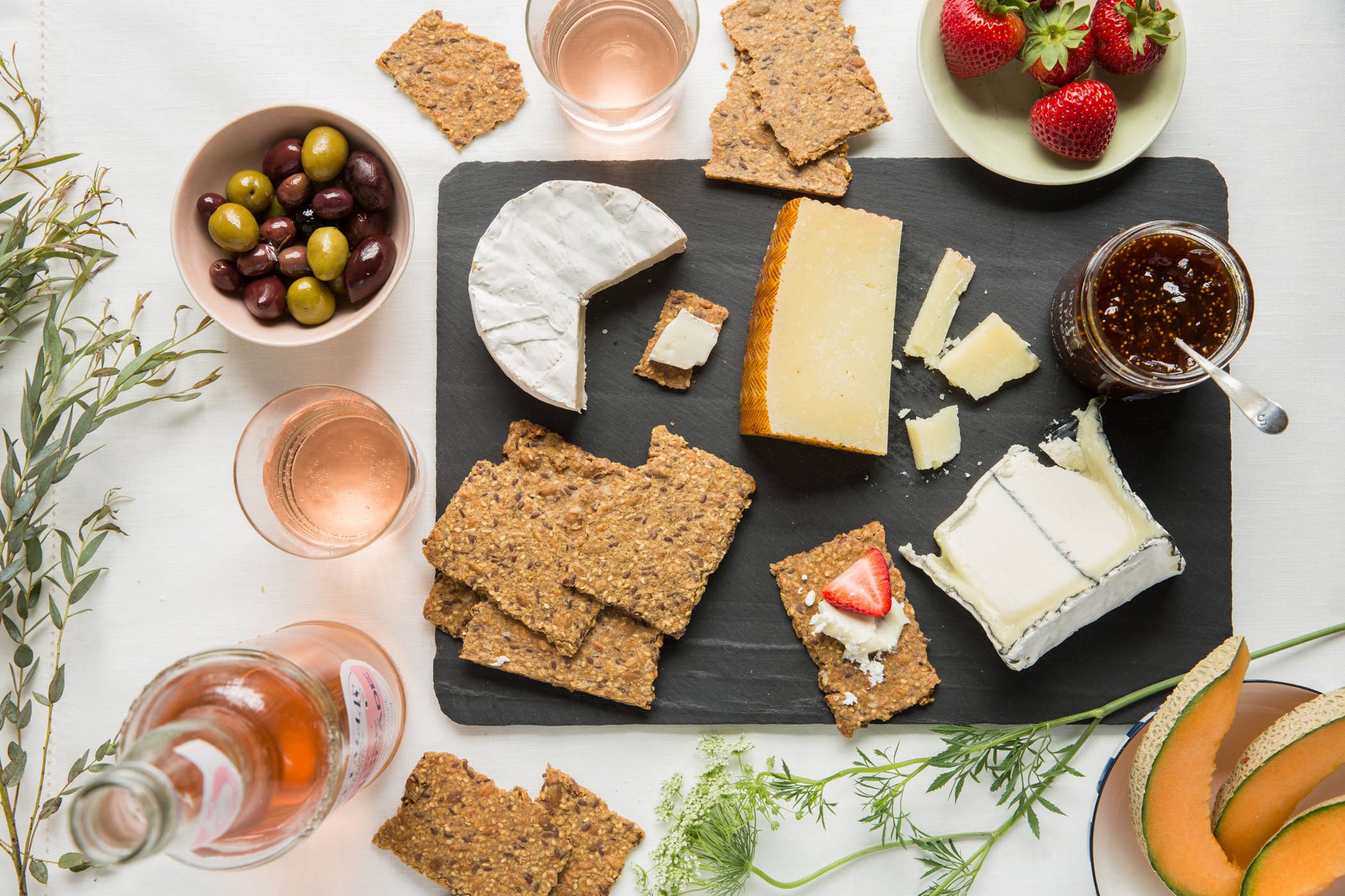 Platter of assorted cheeses, crackers, and fruits with glasses of rose on a white surface.