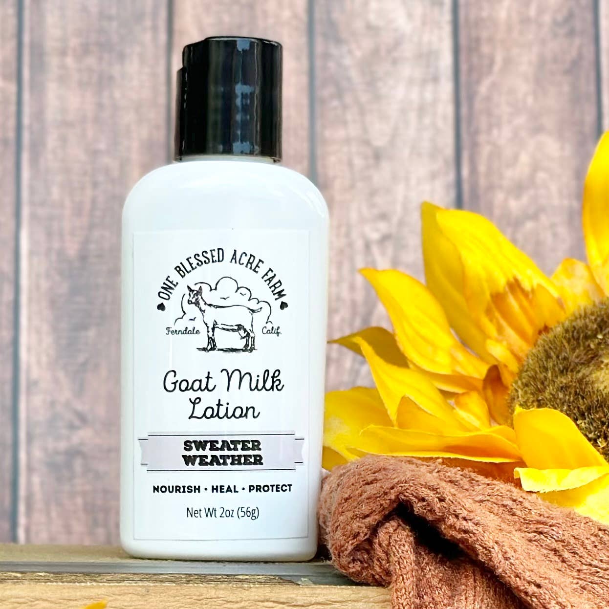 2oz Bottle of Sweater Weather Goat Milk Lotion with a sunflower and towel on a wooden background