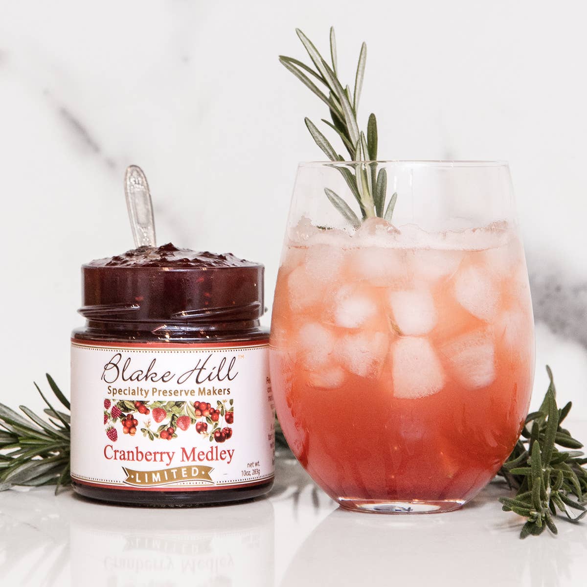 A glass of a red drink with ice and rosemary garnish made with Blake Hill Cranberry Medley Preserves