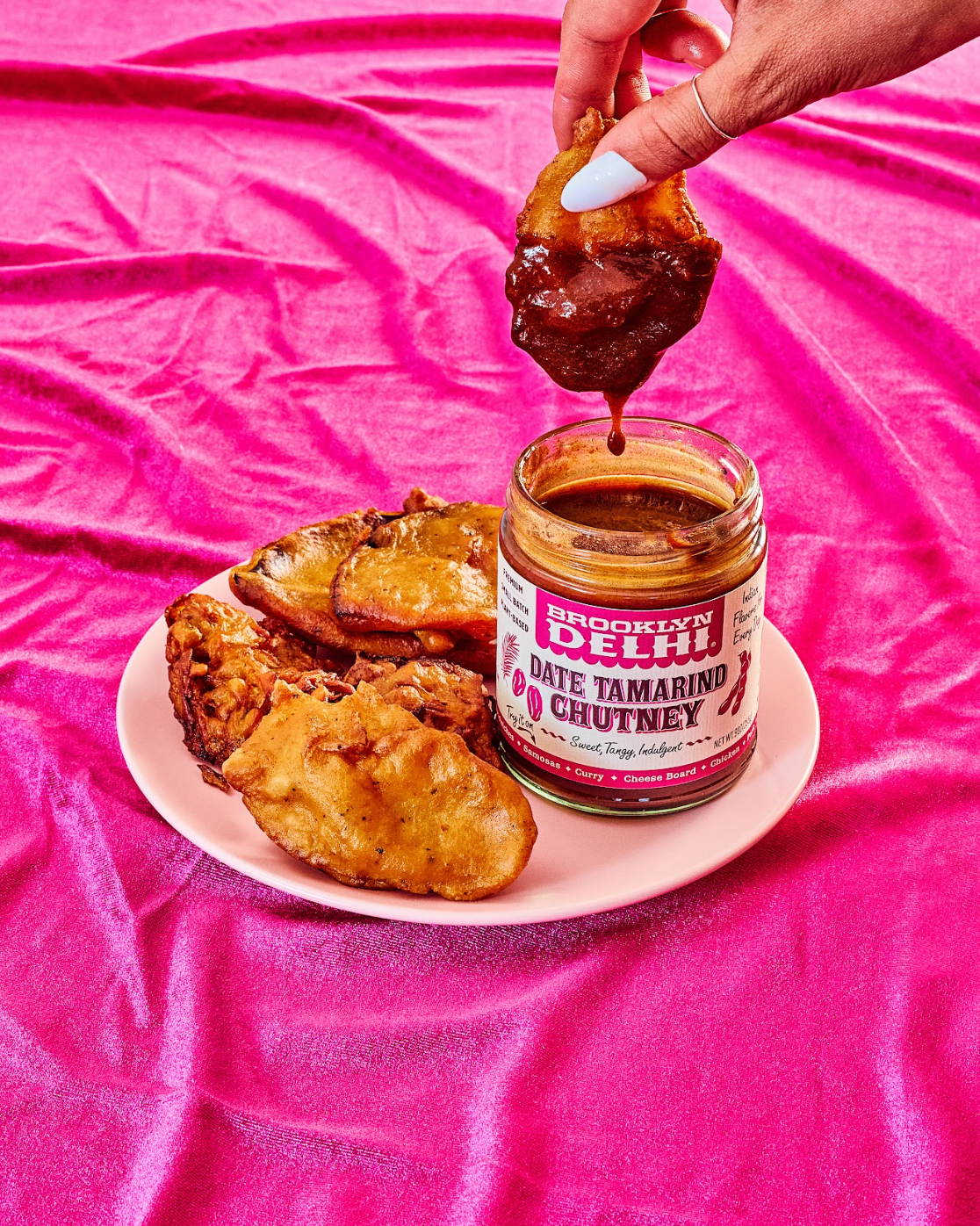 Person dipping fried food into a jar of 'Date Tamarind Chutney' on a pink background
