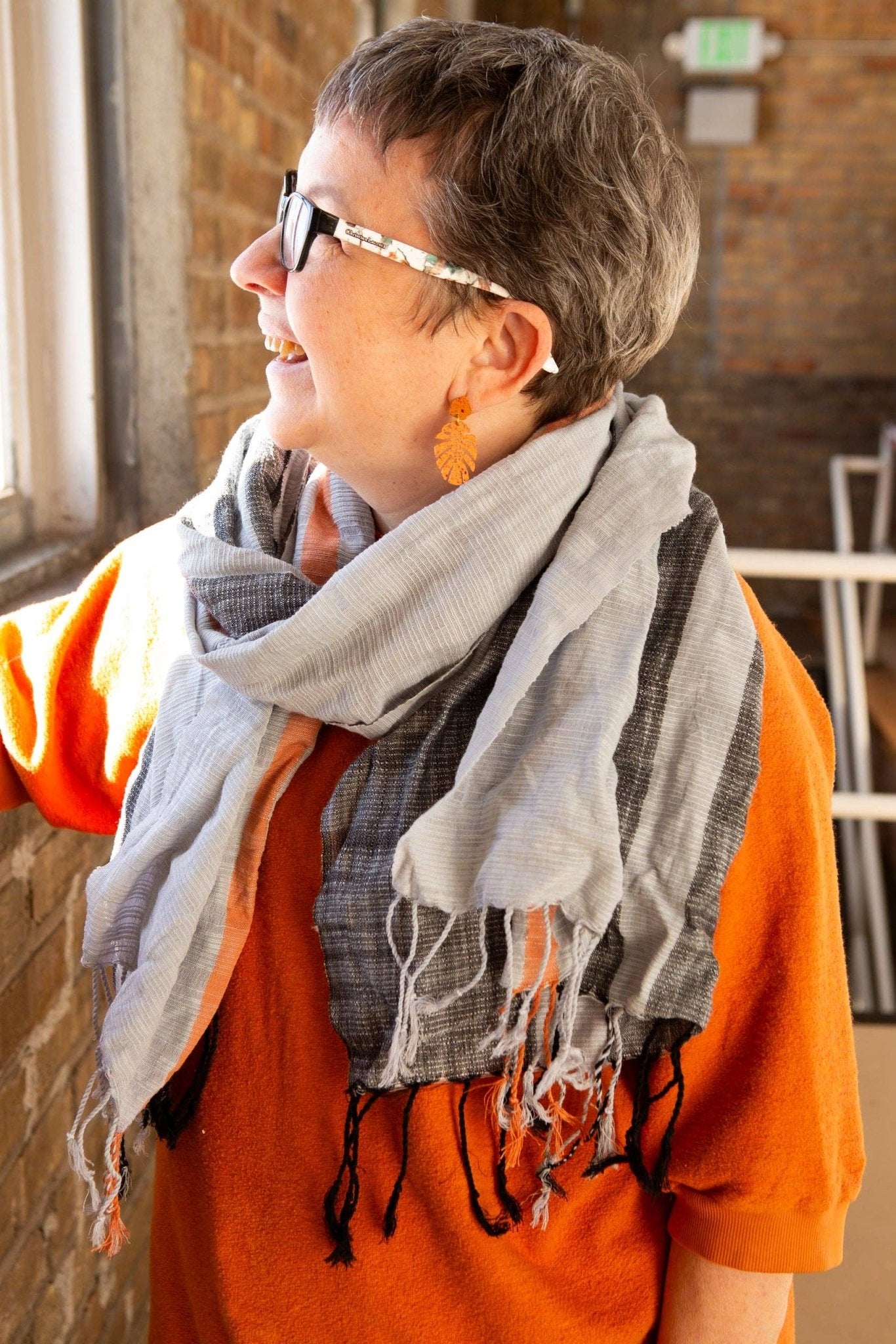 A person wearing an orange sweater and the Graphite Woven Striped Scarf by Fair Anita stands against a brick wall