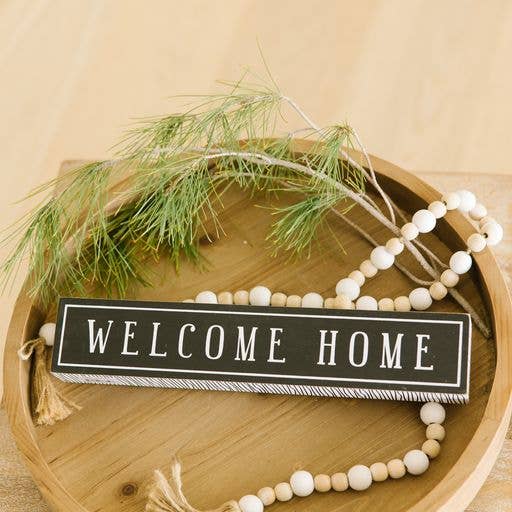 Decorative sign with 'Welcome Home' text on a wooden tray with greenery and beads.