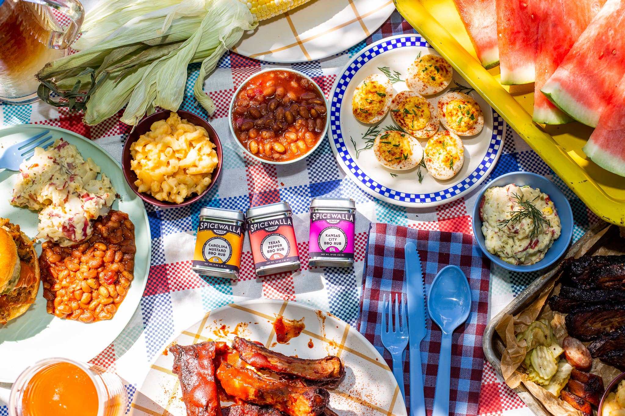 Outdoor picnic setup with plates of food, including burgers, sausages, macaroni and cheese, and Spicewalla Ultimate BBQ Spice Collection. 