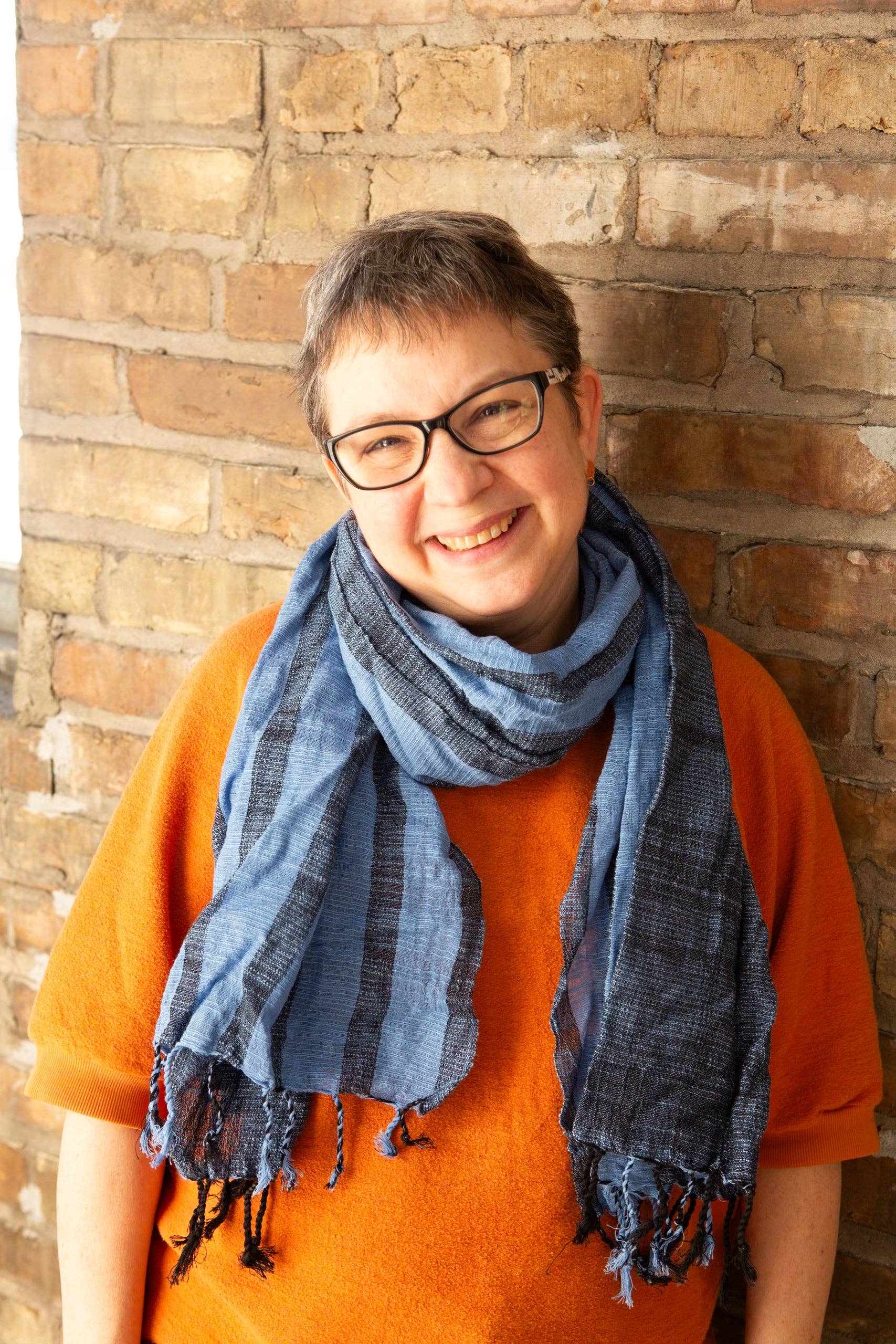 A person wearing an orange shirt and the Stormy Seas Woven Cotton Scarf by Fair Anita stands against a brick wall.