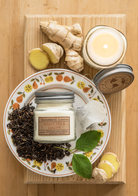White Tea & Ginger Soy Candle by Eco Candle Co on a decorative plate with ginger and tea leaves on a wooden surface.